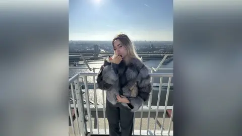 ValerieGracy poses on a balcony wearing a thick grey fur coat and dark trousers against a bright city skyline.