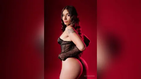 Confident woman in a black sheer bodysuit posing against a moody red background.
