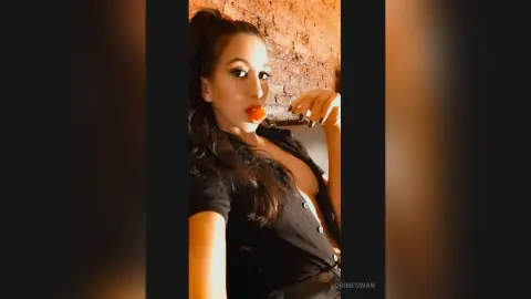 Glamorous woman in a black button-down shirt posing against a rustic brick wall in warm lighting.