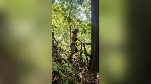 MaddyBrohms stands in a lush, green forest wearing a black tank top and patterned leggings, leaning against a wooden fence near a mossy slope.