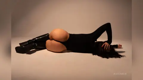 AlesiaNoir — Woman in a black bodysuit and tall leather boots lying on a neutral studio floor in a minimalist pose.