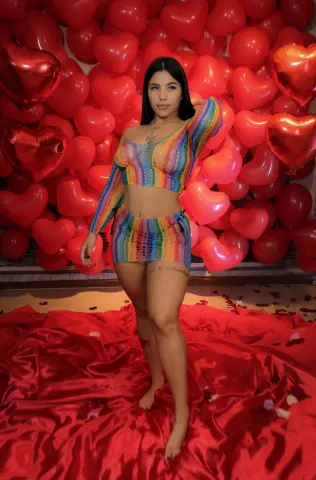channeljonson — Young woman in colorful striped crop top and mini skirt posing against a romantic red heart balloon backdrop.