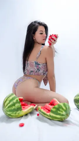 Young woman in floral bikini sitting on white fabric with fresh watermelon slices in a bright studio setting.