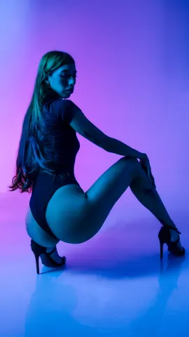 AilynHoney — AilynHoney poses in a black bodysuit and high heels against a blue and purple neon background.