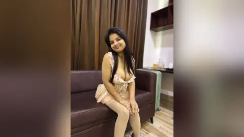 Smiling woman in a lace cream bodysuit sitting on a brown sofa in a cozy living room setting.