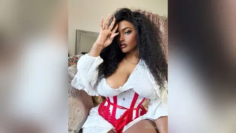 BossyBella — BossyBella poses with her hand to her head, wearing a white long-sleeve top and a red corset, showing off her long, dark curly hair.