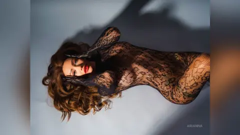 Elegant woman in a sheer black lace bodysuit posing against a soft blue background with dramatic shadows.