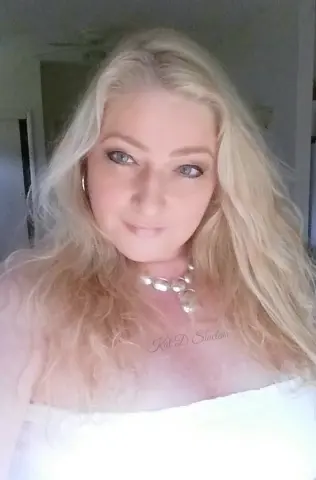 KatDSinclair poses with her long blonde hair down, wearing a white top and a silver necklace.