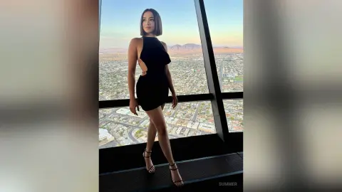 Stylish woman in a black mini dress posing by a skyscraper window overlooking a desert city skyline at sunset.