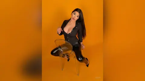 Glamorous woman in a black button-down shirt and leather leggings sitting on a clear chair against an orange background.