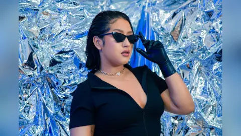 KimRoberth wears black sunglasses and a black zip-up top against a shiny silver foil background.
