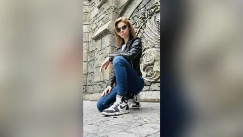 Stylish woman in black leather jacket, blue jeans, and high-top sneakers sitting by a stone wall.