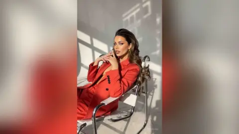 AnaMathers poses in a red blazer and matching bottoms, resting her chin on her hands while sitting in a white chair.