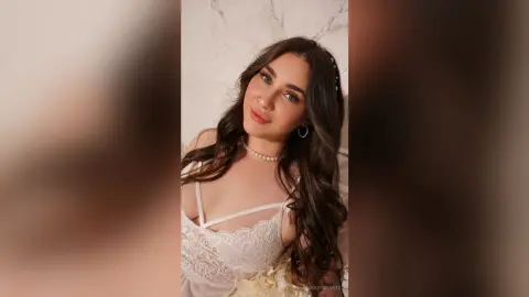 KeiraHarts poses with long wavy brown hair, wearing a white lace bra and a pearl necklace against a light marble wall.