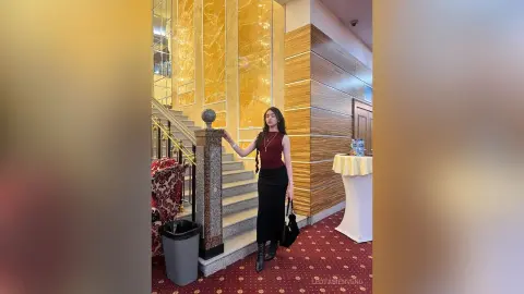 Elegant woman in a burgundy sleeveless top and black maxi skirt standing on a grand marble staircase.