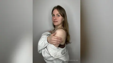 CatherynBuckland poses in a white zip-up hoodie and a brown bra, looking at the camera against a plain grey background.