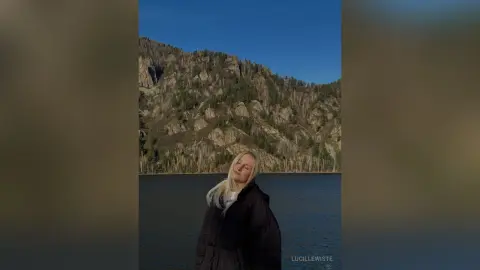 Blonde woman in a black jacket standing by a serene lake with rocky mountains under a clear blue sky.