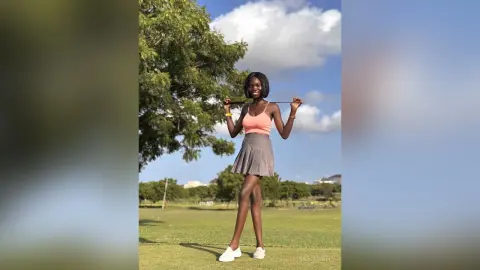 Smiling young woman in a peach tank top and grey skirt standing in a sunny green field under a blue sky.