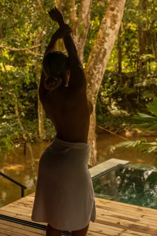 Nude woman in a white towel stretching on a wooden deck by a tropical jungle pool at sunset.