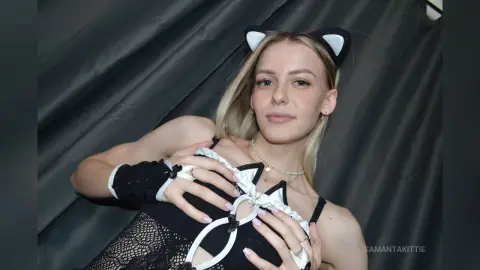 Blonde woman in a black cat costume with white trim and fingerless gloves posing against a dark curtain.