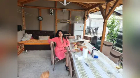 Smiling woman in a pink robe sitting at a dining table in a rustic outdoor patio setting.