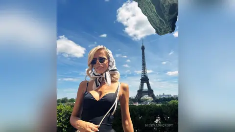 NickyAshcroft poses in a black bodysuit and sunglasses with a white headscarf, with the Eiffel Tower visible behind her under a blue sky.