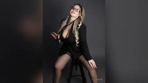 Stylish woman in a black blazer and glasses sitting on a stool against a grey studio background.