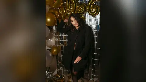 Smiling woman in a black coat celebrating a birthday with gold balloons in a festive indoor setting.