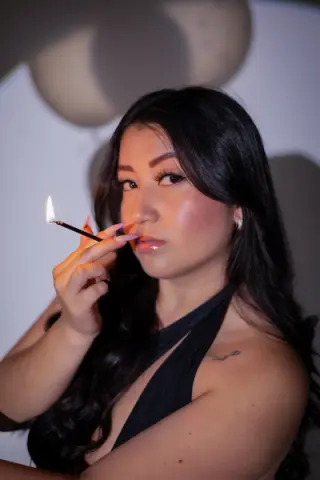 Glamorous woman in a black halter top holding a lit match in a moody, dimly lit setting.