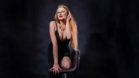 Elegant blonde woman in a black lace dress and glasses posing glamorously on a stool against a dark backdrop.