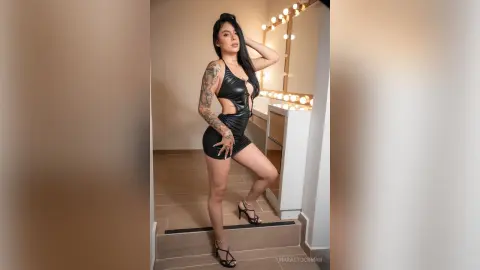 Tattooed woman in a sleek black mini dress posing confidently in a modern, brightly lit dressing room.