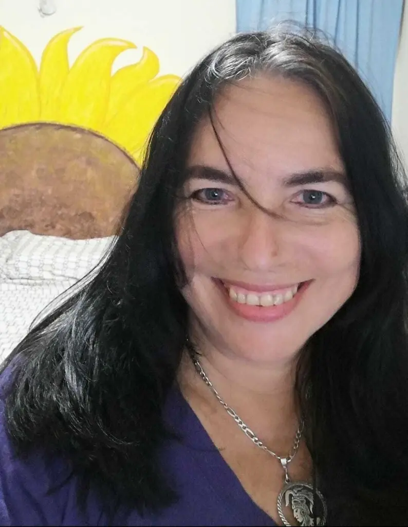 AndreaLoveLatin smiles with long dark hair and glasses, wearing a light blue tank top and a silver pendant necklace.