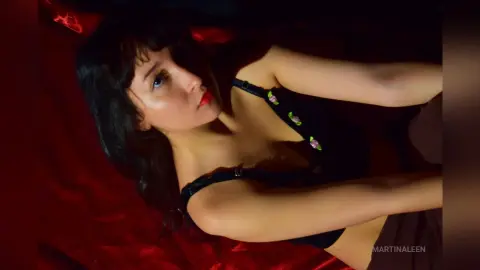 Young woman in a black floral camisole relaxing on red fabric in moody, low-light setting.