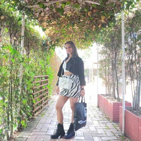 Stylish woman in a black jacket and zebra print skirt standing on a sunlit garden walkway under green vines.