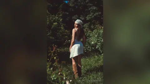 AlessiaCrush poses in a white sundress and headband while standing in a lush green field.