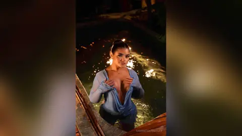 Glamorous woman in a sheer blue top posing by a glowing swimming pool at night.