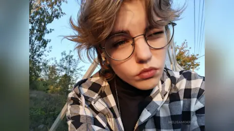 Young woman with messy hair and round glasses wearing a black turtleneck and plaid shirt outdoors.