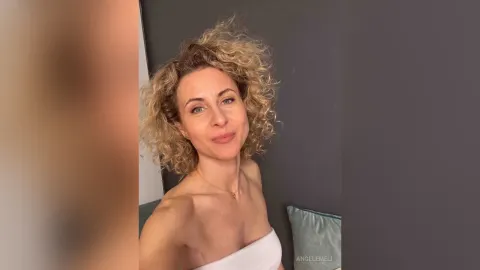 AngelEmeli — AngelEmeli smiles at the camera with her blonde curly hair while wearing a white tube top.
