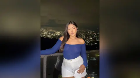 Young woman in a blue off-the-shoulder top and white shorts posing against a glowing city skyline at night.