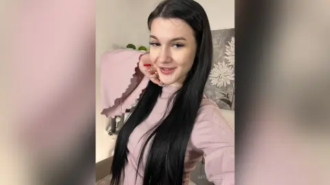 Smiling young woman with long dark hair wearing a cozy pink ribbed sweater in a bright indoor setting.