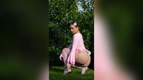Curvy woman in a pink long-sleeve top and sheer tights posing outdoors against a lush green garden background.