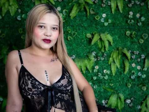 Blonde woman in a black lace camisole posing against a lush green leaf wall background.