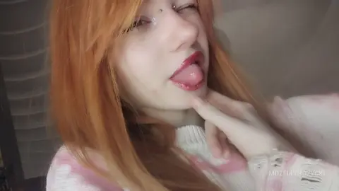 MozelleRozycki licks her lips with her tongue out, showing her red lipstick and nose piercing while wearing a white and pink sweater.