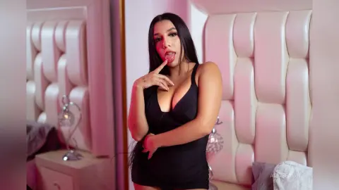 Young woman in a black mini dress posing playfully in a luxury bedroom with a white tufted headboard.