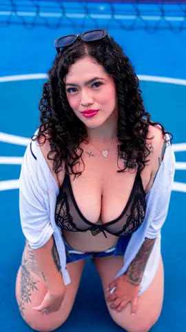 Curvy woman with curly hair in a black lace bra and white shirt posing on a blue tennis court.