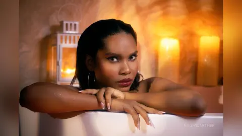 Beautiful woman with dark hair leaning on a white surface against warm glowing candlelight in a cozy setting.