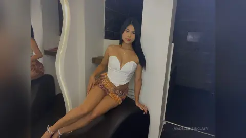 MadeleinSinclair sits on a black bench wearing a white corset top and patterned shorts, posing with long dark hair in a modern room.
