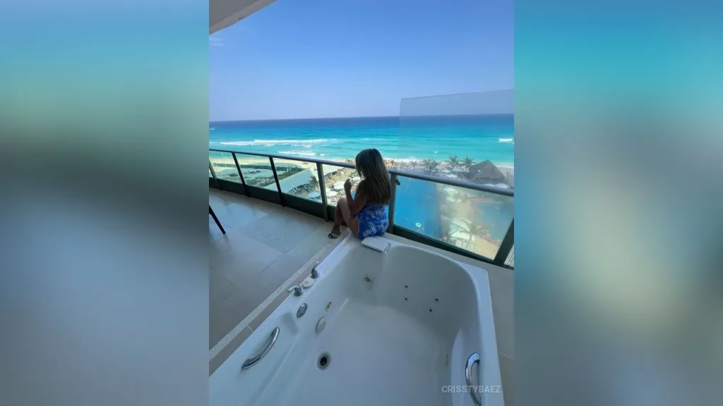 CrisstyBaez — CrisstyBaez sits on a balcony railing overlooking the blue ocean while wearing a blue patterned bikini.