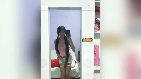 SofiaCorazon poses in a pink tank top and light shorts, holding her phone for a mirror selfie in her bedroom.