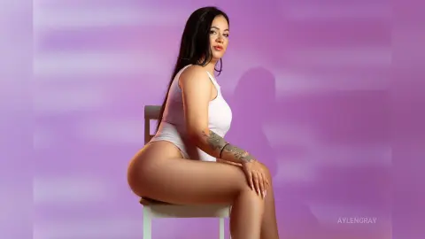 Curvy woman in a white bodysuit sitting on a chair against a soft purple background with a serene mood.